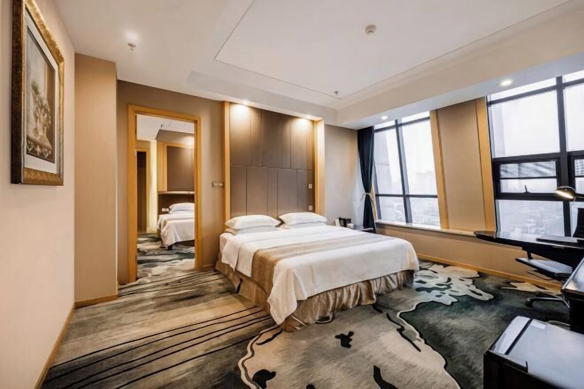 A hotel room with a large bed, a desk with a computer, and a view of the city through large windows.