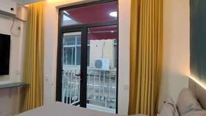 Ein Hotelzimmer mit einem Bett, einem an der Wand befestigten Fernseher, einem Fenster mit gelben Vorhängen und Blick auf Gartenmöbel.