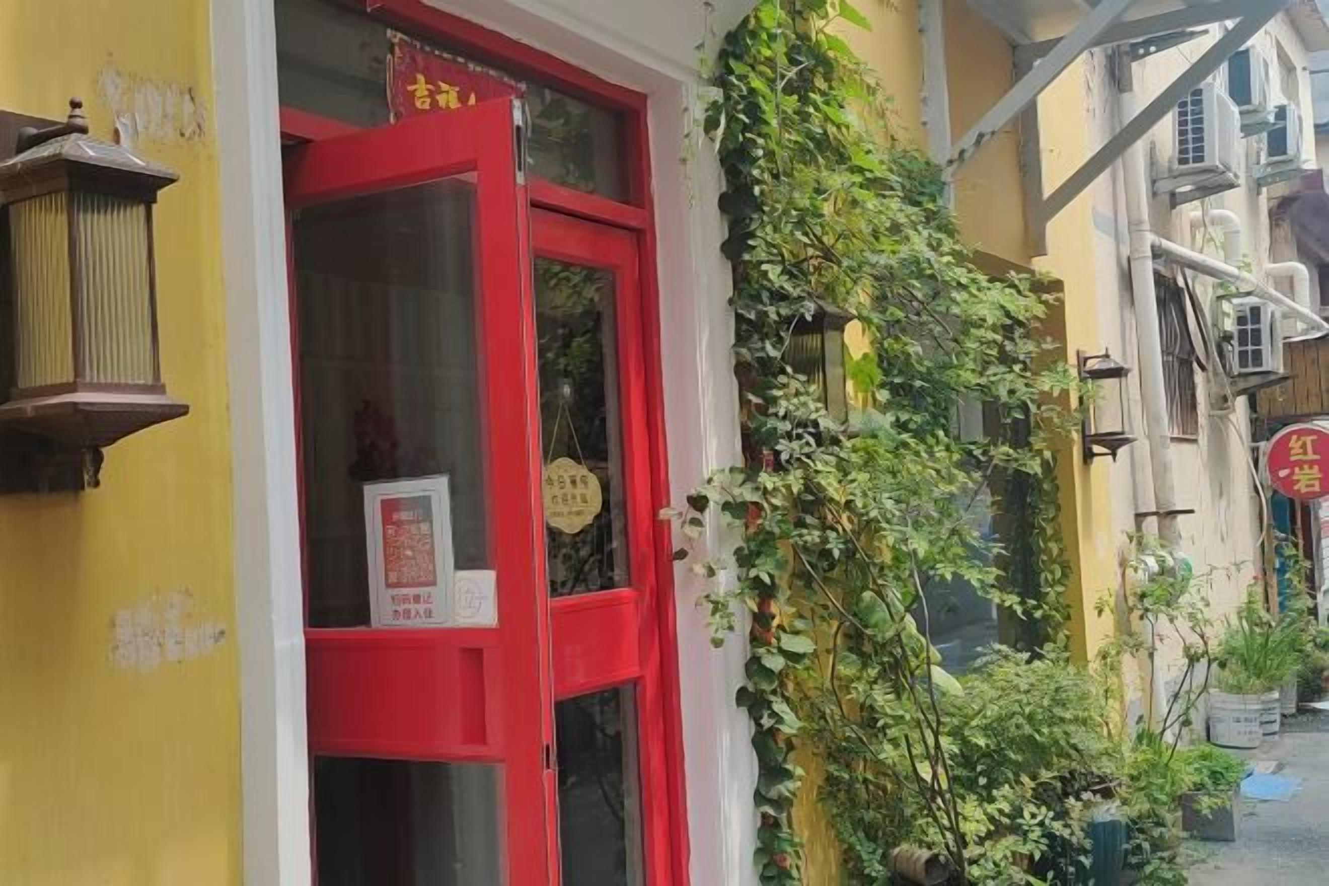 A building with a red door and a sign above it. There is greenery growing on the wall next to the building.