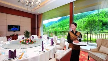 Een restaurantgedeelte in een hotel met een ronde tafel gedekt voor een maaltijd, een televisiescherm en uitzicht op een groen landschap door grote ramen.