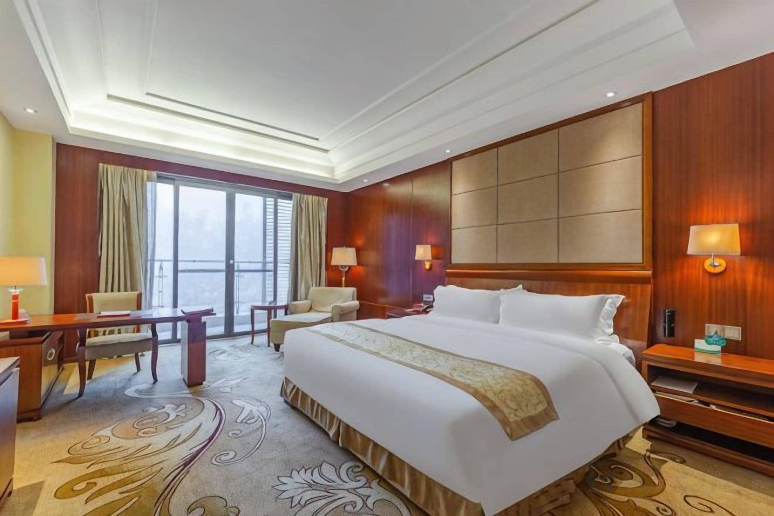Une chambre d’hôtel avec un grand lit, deux tables de chevet, un bureau avec une chaise, un canapé et une grande fenêtre avec des rideaux.