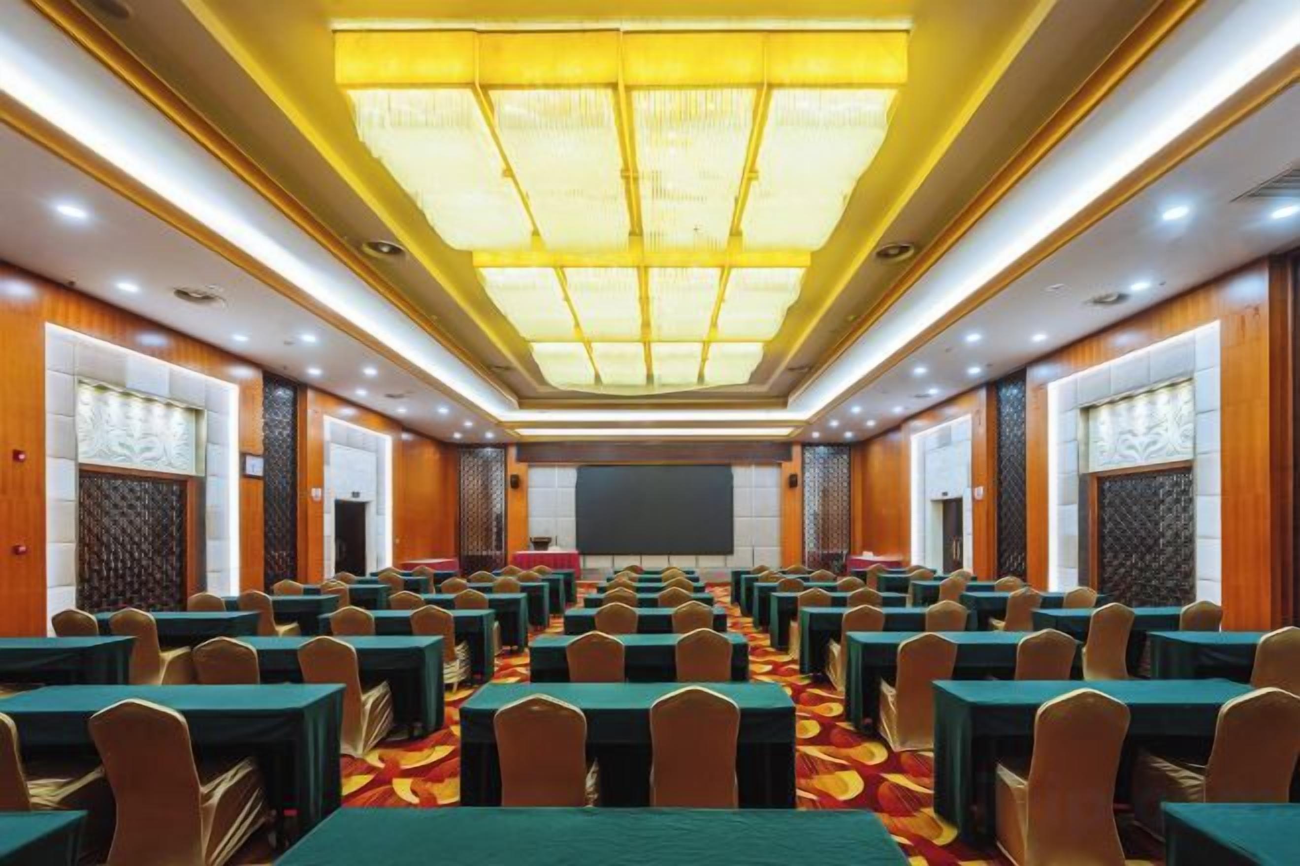 Un salón de conferencias con filas de mesas y sillas, una pantalla grande al frente y una alfombra estampada.