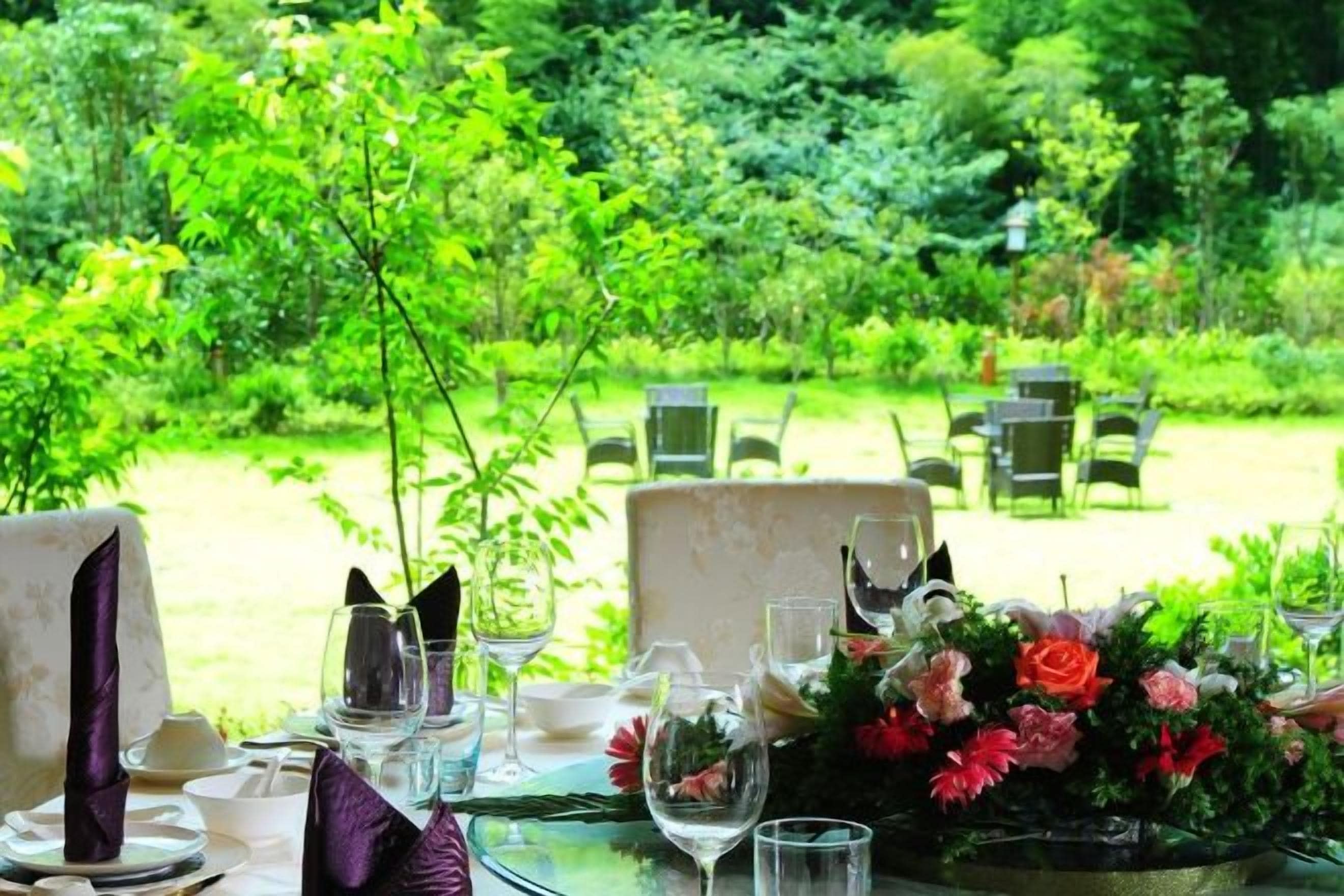 An outdoor dining area with tables set for two, surrounded by lush greenery.