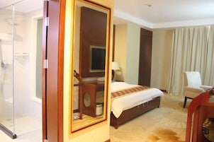 A hotel room with a bed, a desk, a chair, a mirror, and a shower area.