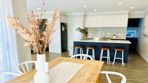 A modern kitchen with white cabinets, a wooden dining table with a white vase of dried flowers, and white chairs. - Serenity by the Bay - Whole House in Vincentia, Jervis Bay (Vincentia)