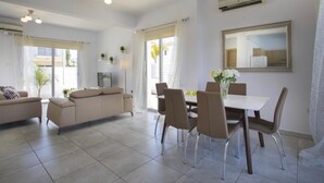 Amplia sala de estar con sofá, mesa de comedor y sillas. Hay plantas en macetas y un televisor sobre un mueble.