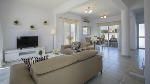 Un salón amplio con un sofá beige grande, un televisor de pantalla plana y una zona de comedor con mesa y sillas.