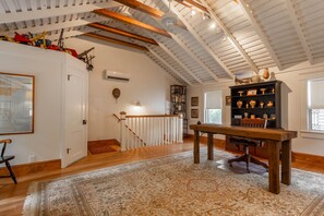 Une pièce spacieuse avec des poutres en bois au plafond, un escalier doté d’une rampe blanche, une table en bois et une bibliothèque remplie d’objets décoratifs.