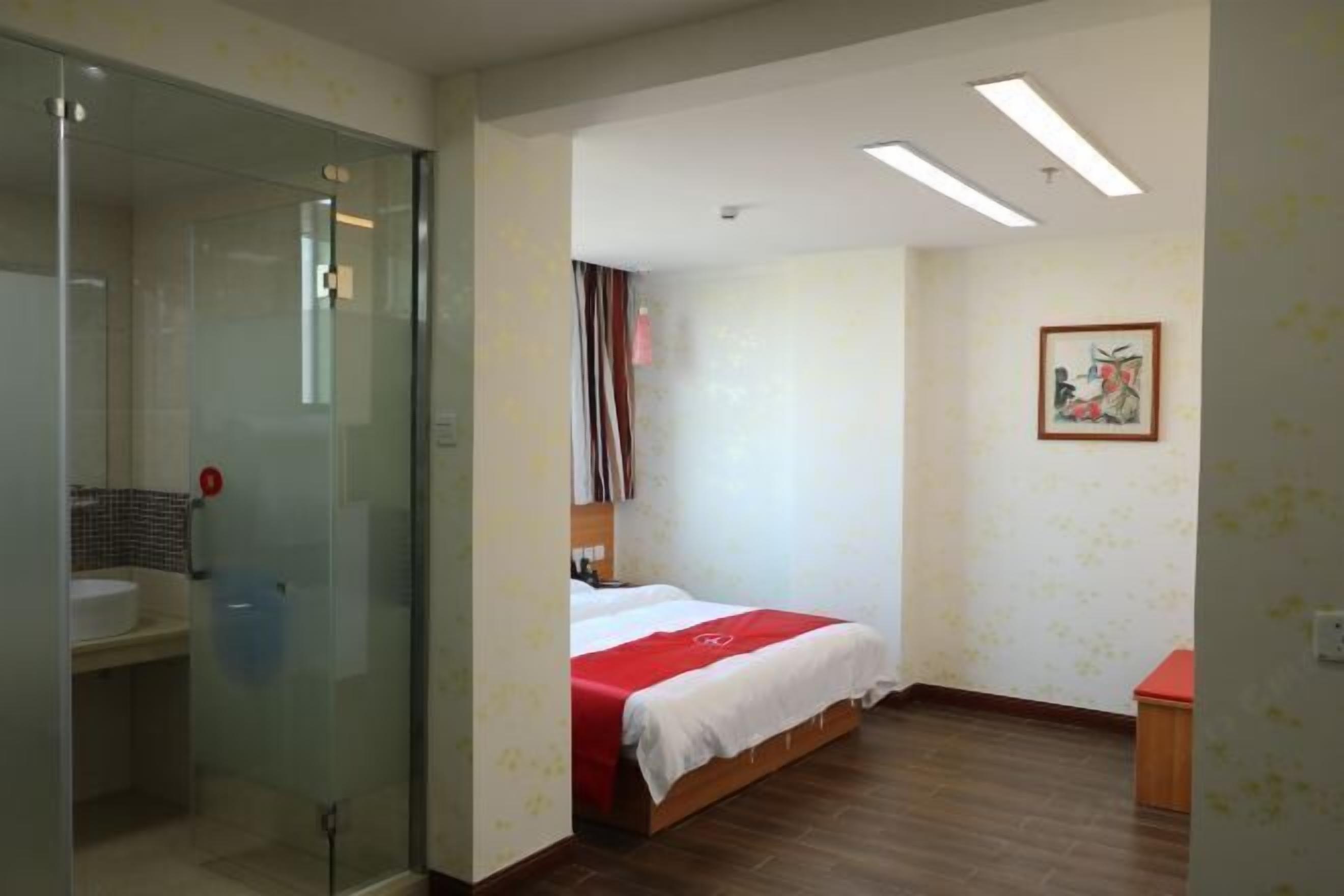 A hotel room with a glass partition separating the bathroom, a bed with a red and white bedspread, and a small wooden nightstand.