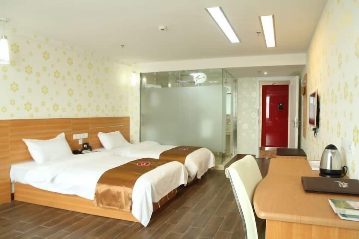 A hotel room with two beds, a wooden headboard, a desk with a kettle, and a red door.