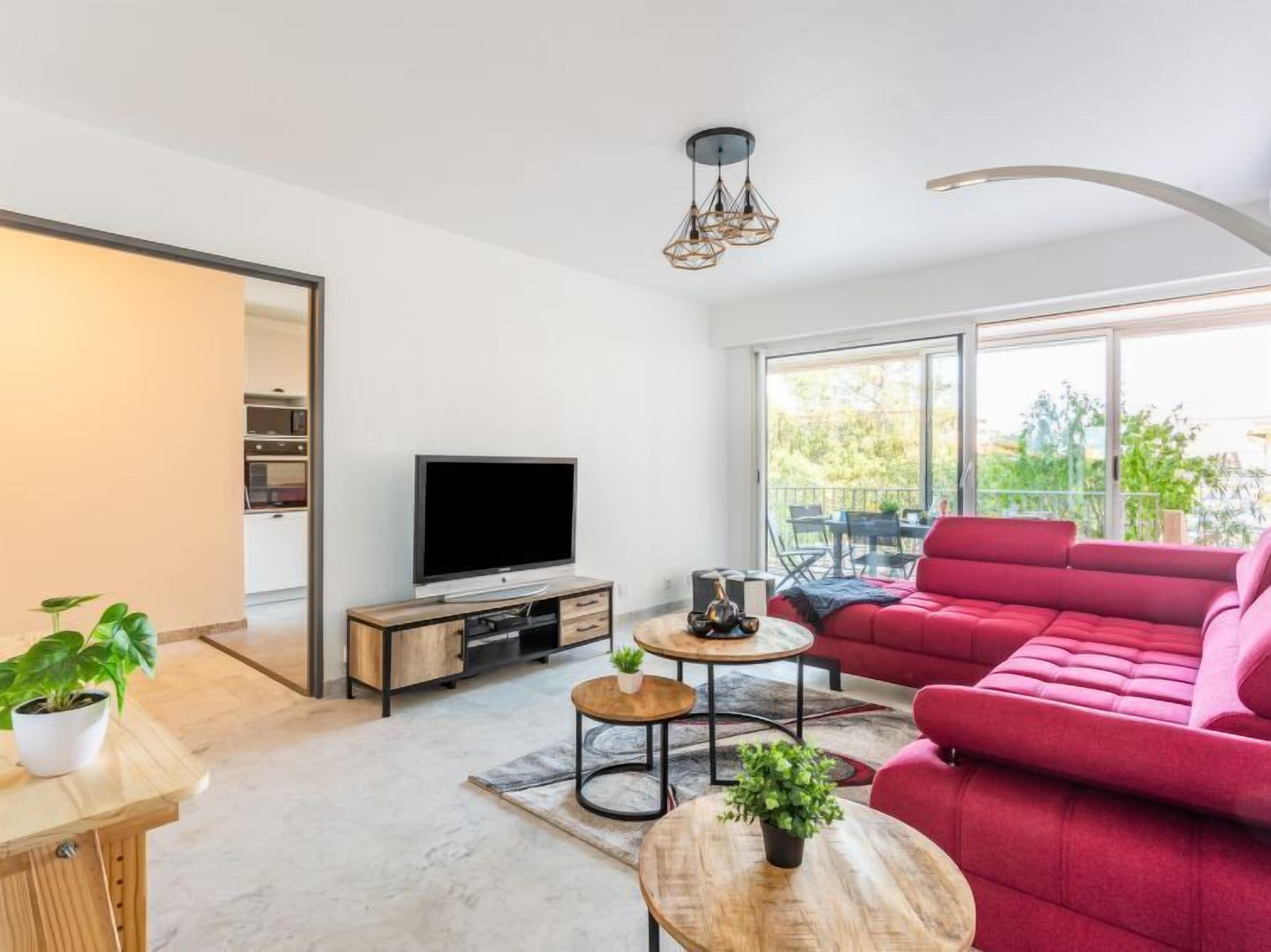 A modern living room with a large window, a red sofa, a TV, and a dining area with a table and chairs.