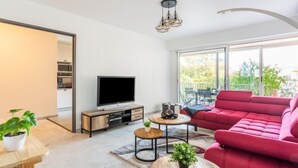 A modern living room with a large window, a red sofa, a TV, and a dining area with a table and chairs. - Le Phenicia by Interhome (Saint Laurent du Var)