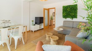 Een moderne woonkamer met een grijze bank, een eettafel met witte stoelen, een televisie op een witte kast, een hangplant en een houten vloer.