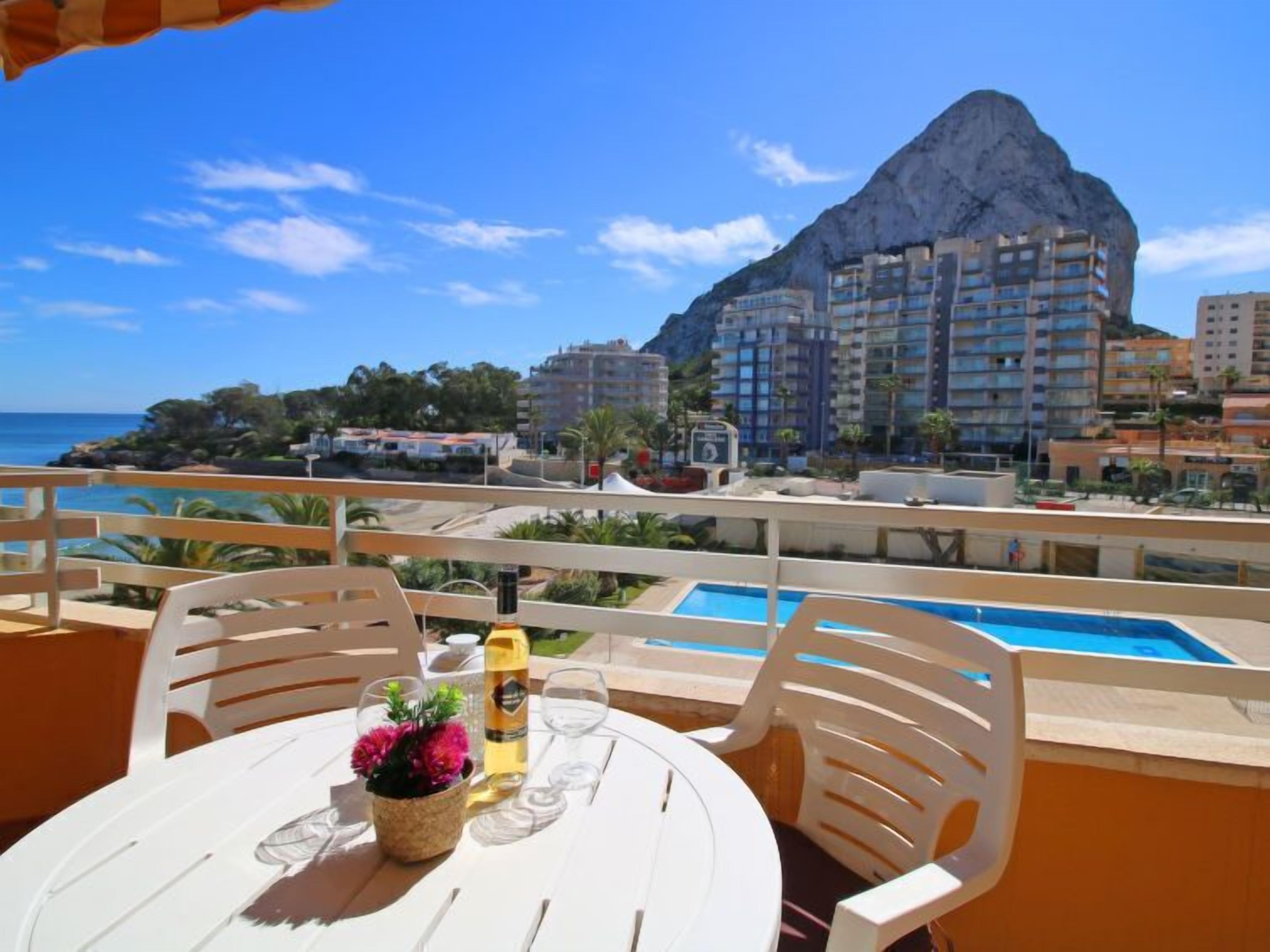 A balcony with a pool, white chairs, and a table with a bottle and flowers.