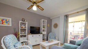 Ein Wohnzimmer mit Fernseher, weißen Möbeln, Deckenventilator und Blick ins Freie.