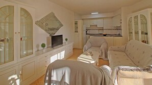 Ein Wohnzimmer mit einer Couch, einem Couchtisch und einem Fernseher. Im Hintergrund ist eine Küche mit Schränken und einem Herd zu sehen.