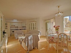 Ampla sala de estar com sofá, mesa de jantar e cadeiras. Cozinha visível ao fundo, com armários e pia.