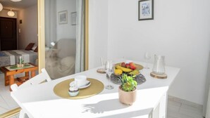 Un coin repas avec une table blanche dressée pour deux personnes, agrémenté d’un panier de fruits, d’un verre et d’une petite plante.