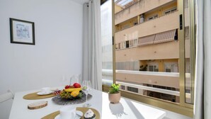 Une chambre moderne avec une table à manger dressée pour deux personnes, une petite plante en pot et un balcon offrant une vue sur un immeuble  à plusieurs étages.