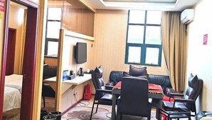 Quarto de hotel compacto com cama, escrivaninha e cadeira, sofá com almofadas, televisão e janela com cortinas.