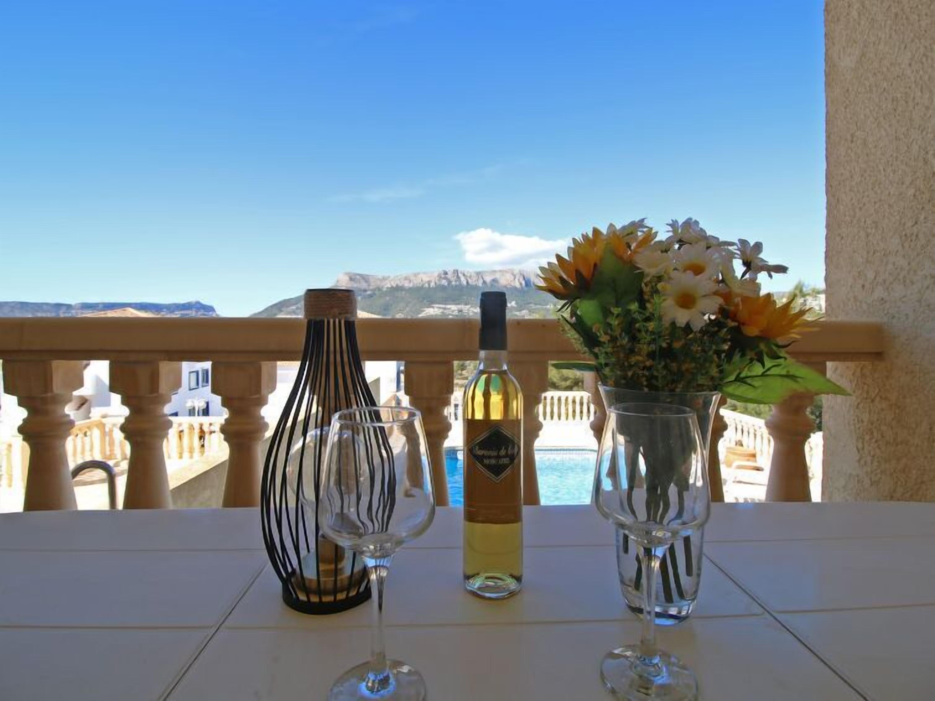 Une table avec une bouteille de vin, deux verres à vin et un vase de fleurs, le tout placé sur un balcon donnant sur un paysage magnifique.