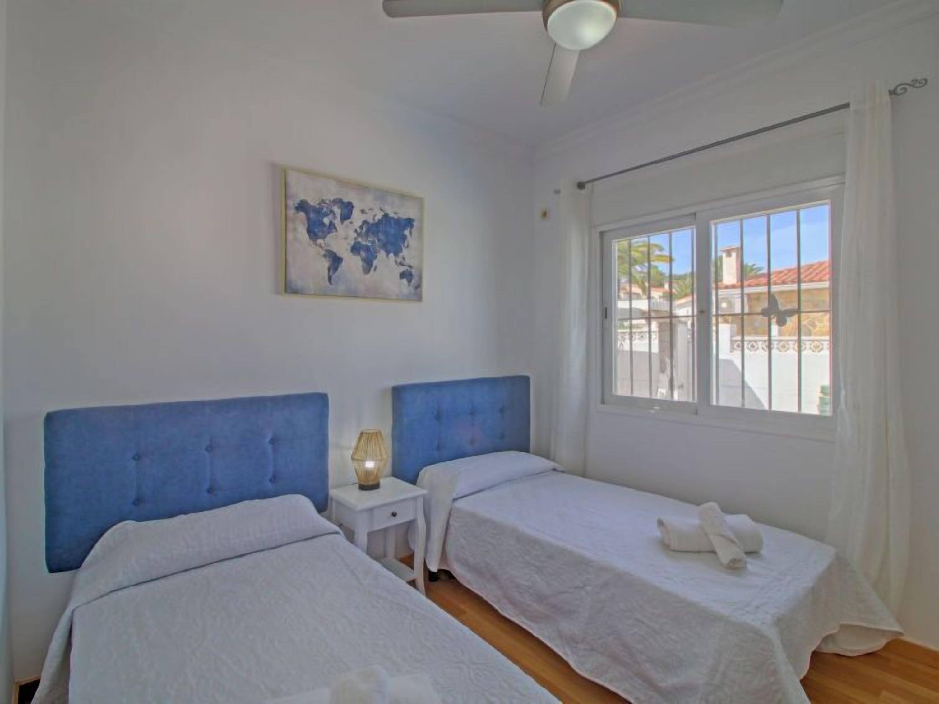 A room with two beds, a small bedside table with a lamp, a world map on the wall, and a window with a view of buildings outside.
