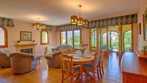 A spacious dining area with a wooden table and chairs, a fireplace, and large windows offering views of greenery.