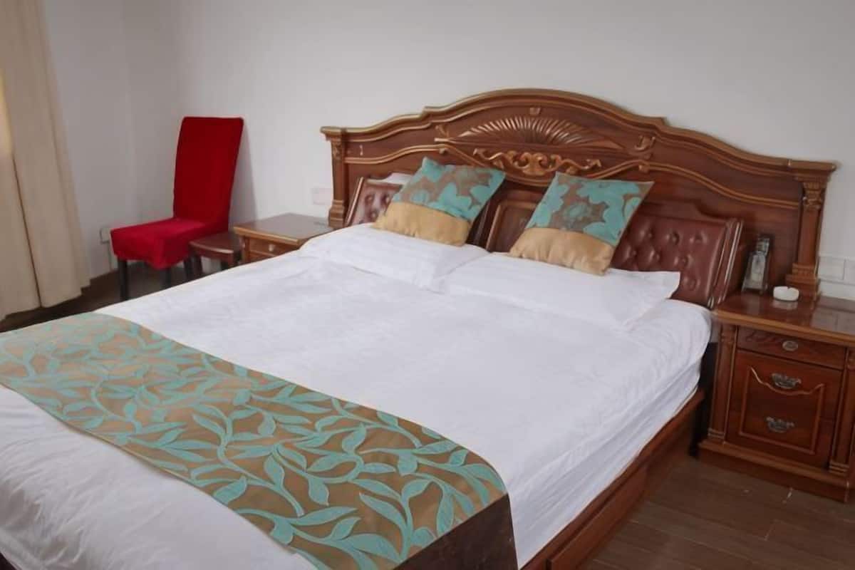 Quarto de hotel com cama de madeira, duas almofadas, uma cadeira vermelha, mesa de cabeceira e um criado-mudo.