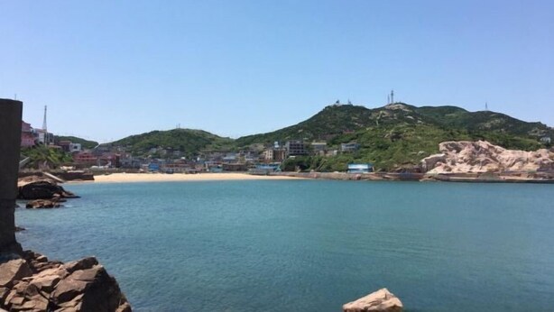 A coastal scene with a sandy beach, calm water, and a hill with buildings in the background. - Shengsi Haiwang Mingzhu Homestay (Shengsi)