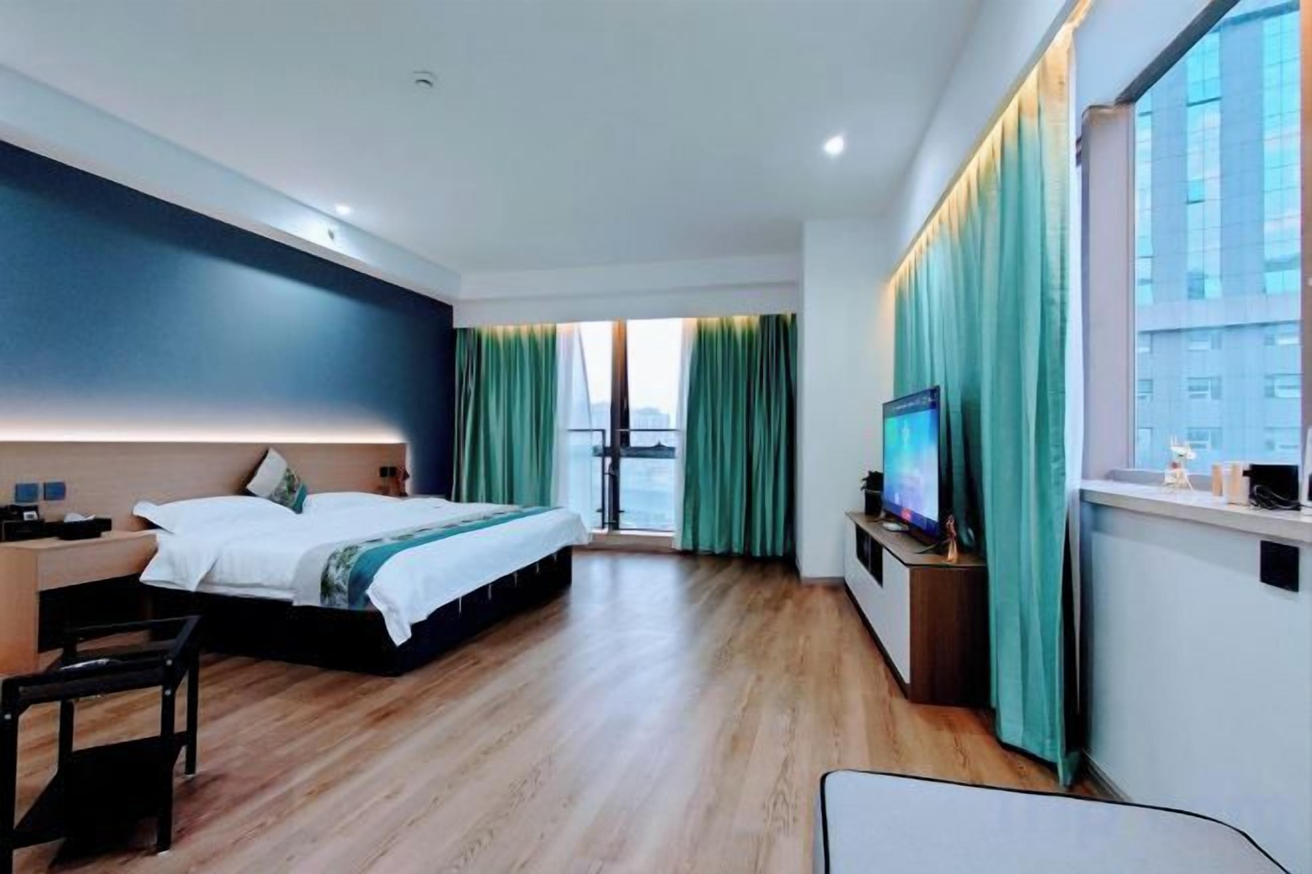 A modern hotel room with a bed, a desk, a chair, a TV, and a view of the city through large windows.