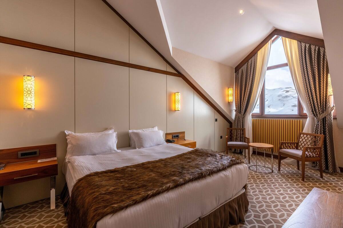 A hotel room with a large bed, a desk, a chair, and a view of a snowy landscape through a window.