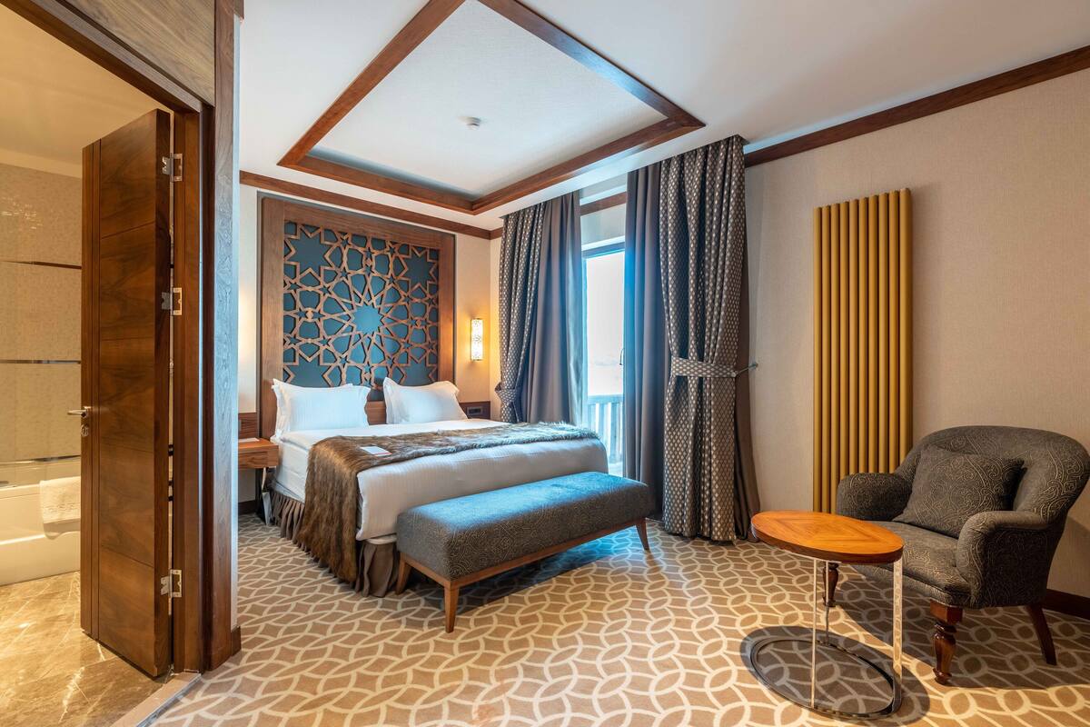 A hotel room with a large bed, a bench, a small table, and a view of the outdoors.