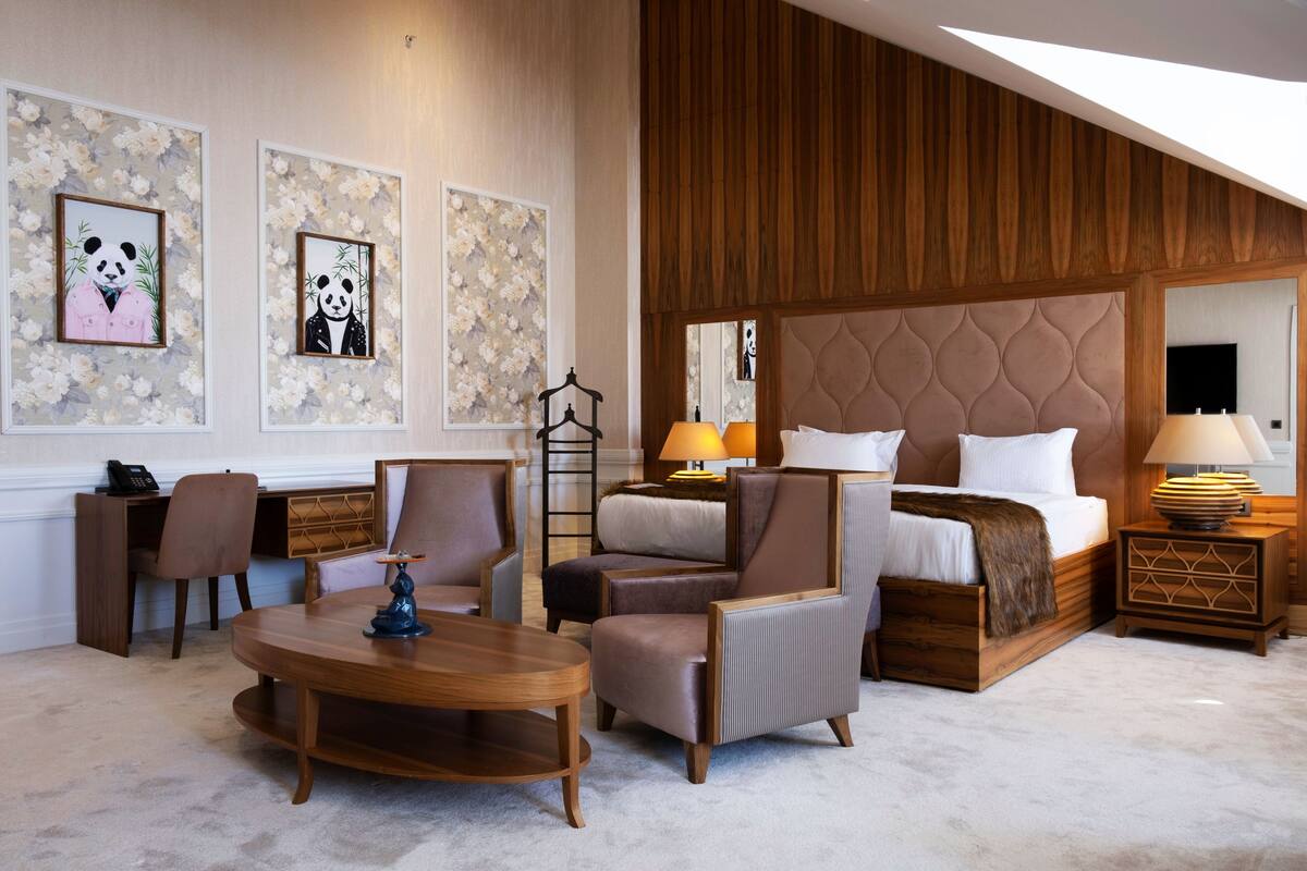 A spacious hotel room with a large bed, a sitting area with a coffee table, a desk, and two chairs. There are framed artworks on the wall and a wooden panel design on the ceiling.