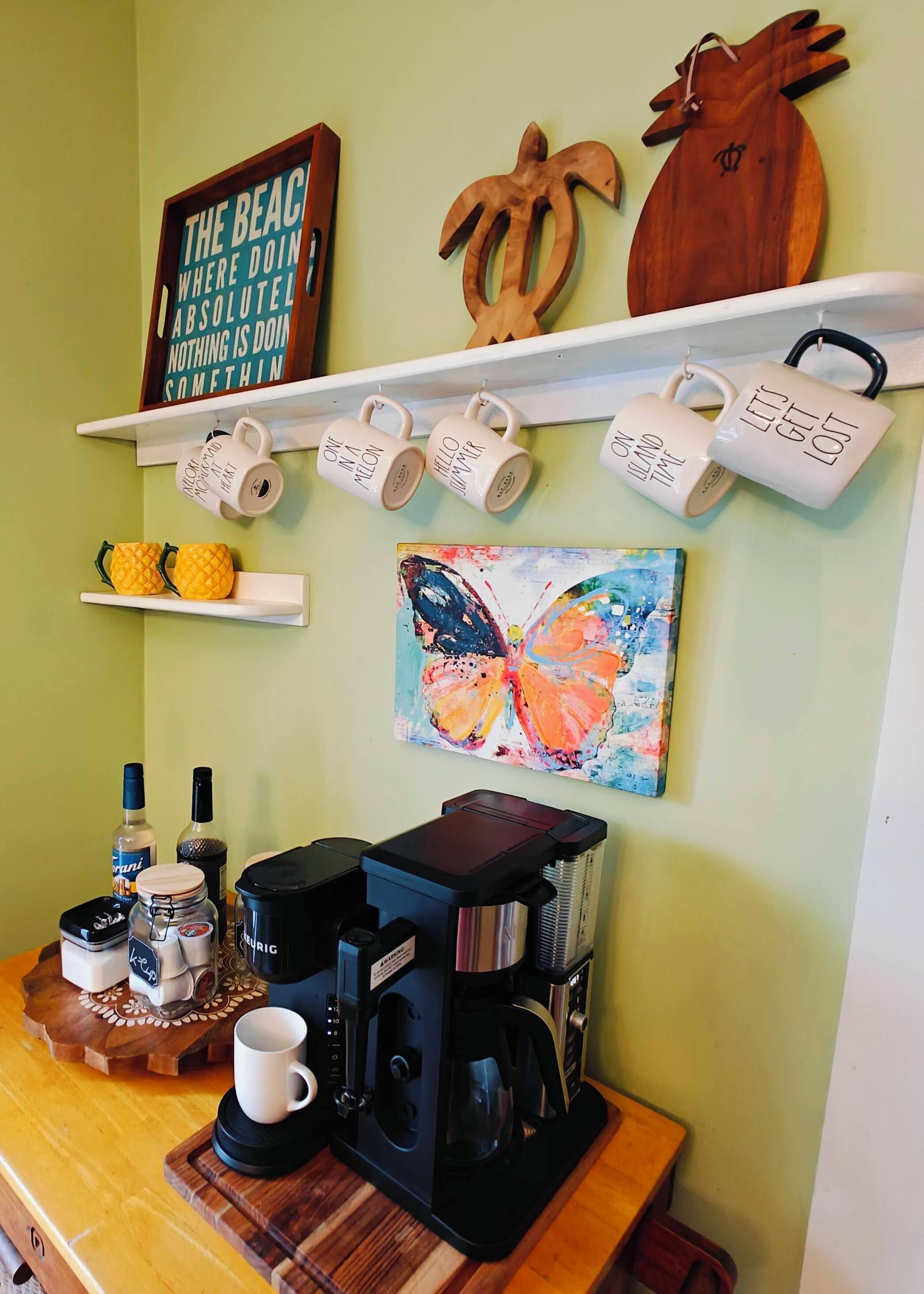 A kitchen area with a coffee maker, mugs, and a shelf with decorative items.