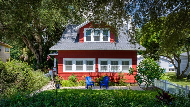 Ein rotes Haus mit weißem Zaun, zwei blaue Stühle im Vorgarten und ein großer Baum zur Linke.