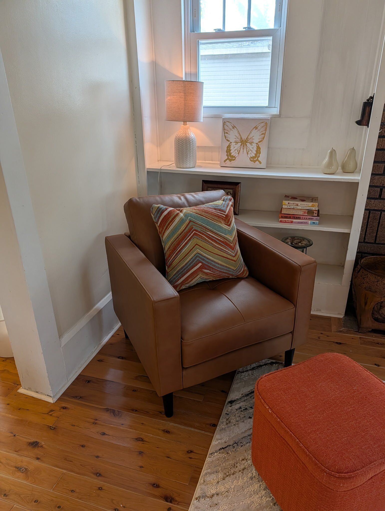 Een gezellige woonkamer met een bruine leren bank, een kleurrijk gestreept kussen, een klein oranje pouf, een witte lamp en een raam met jaloezieën.