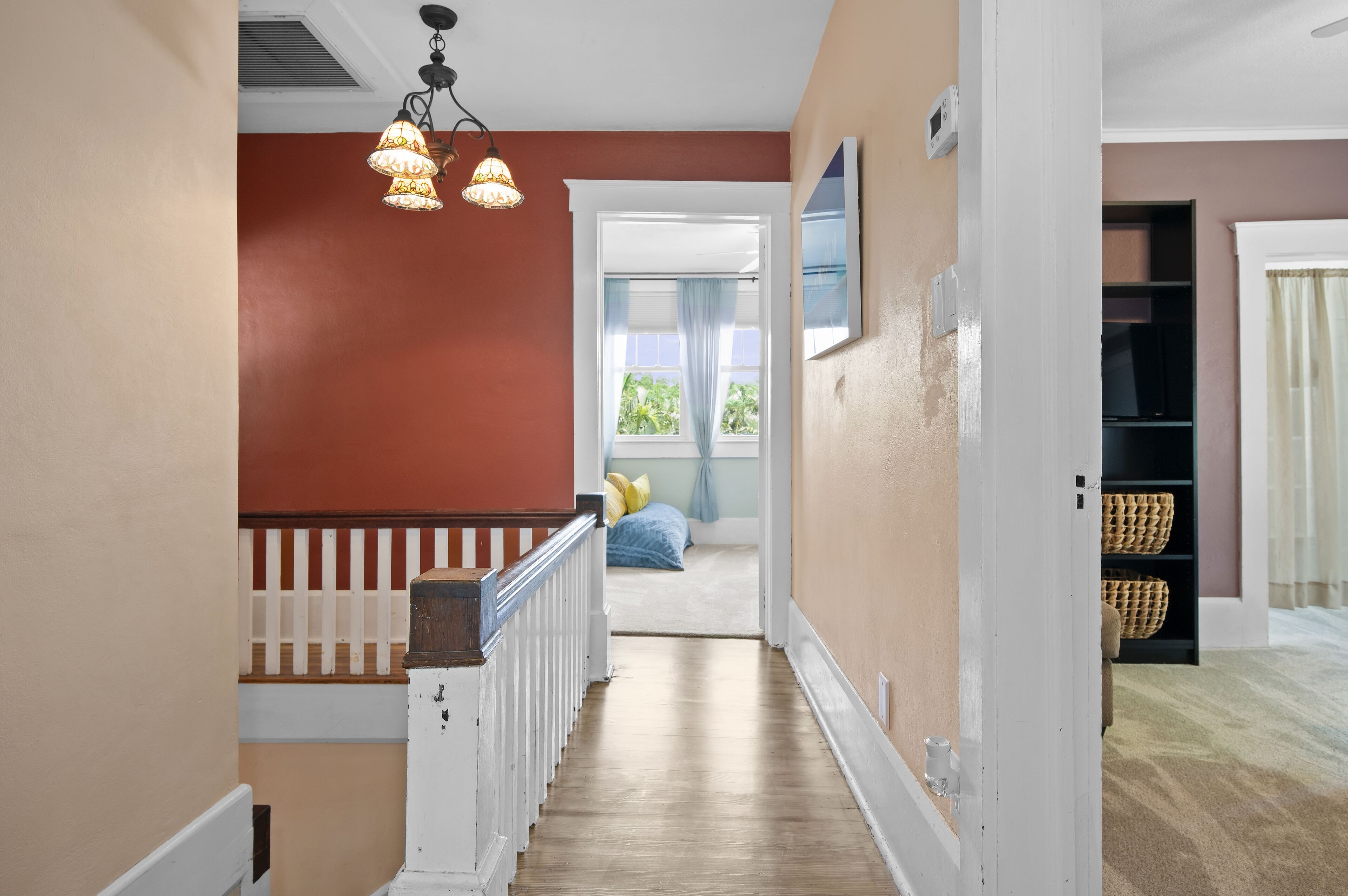 A hallway with a wooden staircase, a ceiling light fixture, and a view into a bedroom with a bed and curtains.
