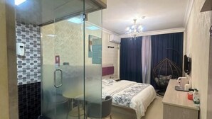 Habitació d'hotel amb una dutxa de vidre, un llit amb un estovat estampat, una taula de nit amb una làmpada i una cadira.