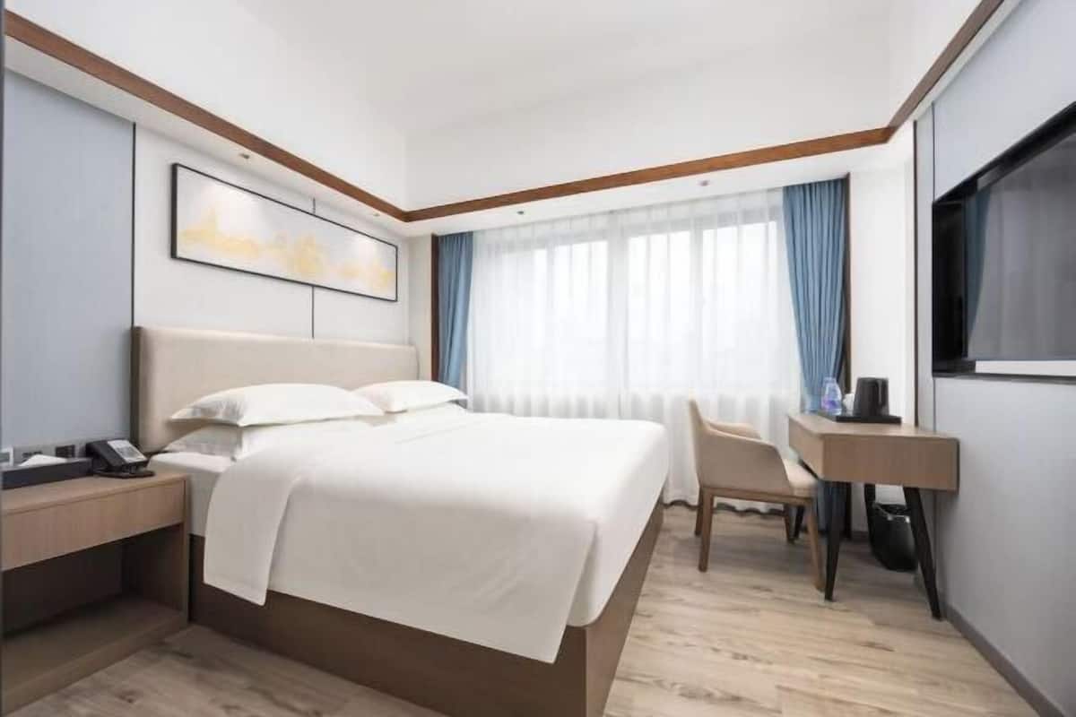 A hotel room with a bed, bedside table, chair, desk, and a TV.