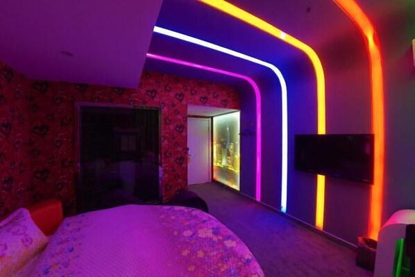 A modern hotel room with a curved ceiling, neon lighting, a bed, a TV, and a hallway with a glass door leading to a bathroom.
