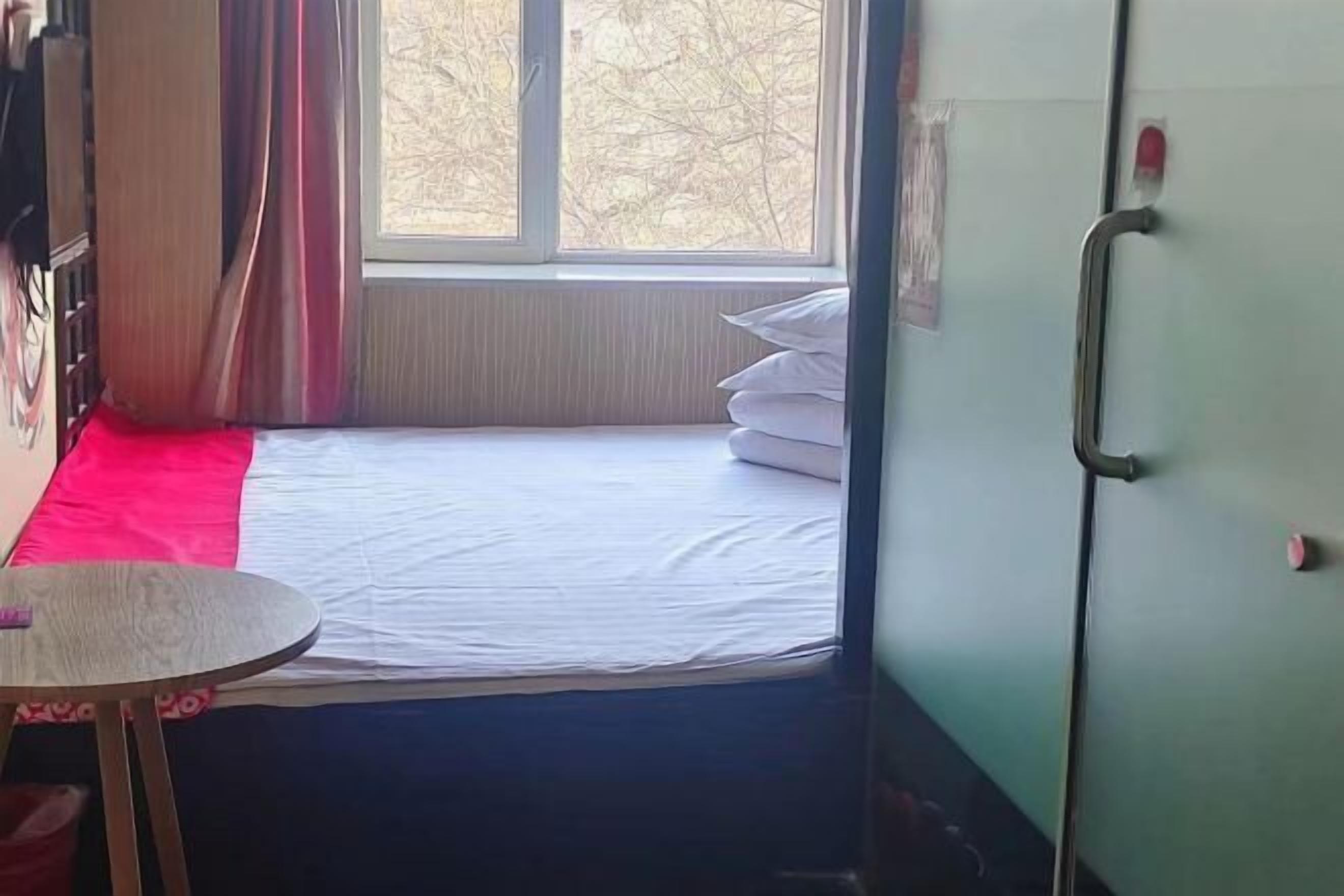 A small room with a single bed, a window with curtains, a small table, and a door with a handle.