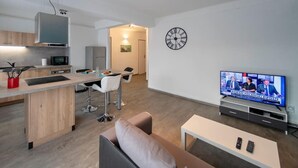 Een modern appartement met keuken, eethoek en zithoek. Er is een televisie, een klok en een bank.