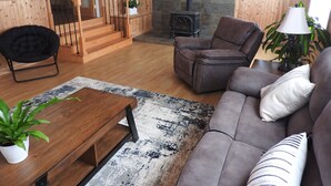 Ein Wohnzimmer mit einer Couch, einem Sessel, einem Couchtisch aus Holz und einem Teppich. Eine Holztreppe mit Geländer und ein Blumentopf sind ebenfalls zu sehen.