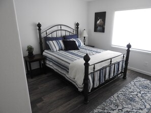 A neatly made bed with a striped bedspread and pillows, a bedside table with a lamp, a potted plant, and a large window with a view of the outdoors. - Coastal Charm: SW Oregon home on quiet 2+ acres, 5min. to the beach! (Gold Beach)
