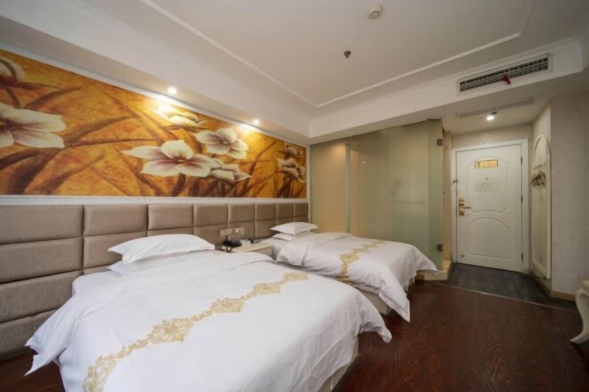 A hotel room with two beds, a large floral wall mural, a door with a sign, and a ceiling vent.