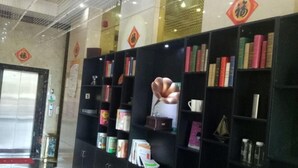 Un vestíbulo con una estantería negra llena de libros y revistas, un jarrón decorativo grande y un letrero rojo y dorado en la pared.