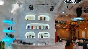 Zona de bar con estantes iluminados mostrando botellas, una recepción y un área de descanso con mesas y sillas redondas.