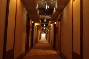 Un long couloir étroit, typique des hôtels, avec des portes des deux côtés, des luminaires au plafond et un sol recouvert de moquette.