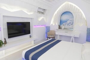 Habitación de hotel con un televisor de pantalla plana grande, una cama con colcha azul y blanca, un escritorio con silla y un espejo montado en la pared.