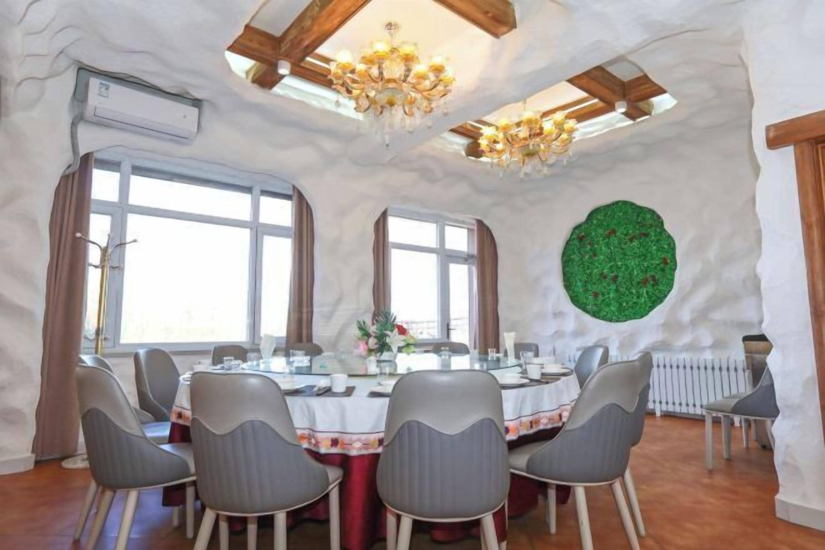 A dining room with a round table, grey chairs, chandeliers, and a green wall decoration.